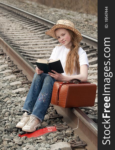 The young girl with a fair hair in a white scarf on a head with a brown sac sits on railway rails in expectation of a train and reads the book. The young girl with a fair hair in a white scarf on a head with a brown sac sits on railway rails in expectation of a train and reads the book