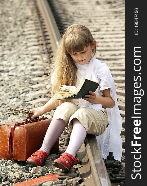 The young girl with a fair hair in a white scarf on a head  with a brown sac sits on railway rails in expectation of a train and reads the book. The young girl with a fair hair in a white scarf on a head  with a brown sac sits on railway rails in expectation of a train and reads the book