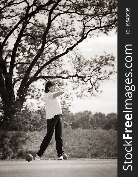 Active senior female golf player swinging golf club to tee off ball with beautiful tree in background, black & white conversion. Active senior female golf player swinging golf club to tee off ball with beautiful tree in background, black & white conversion.