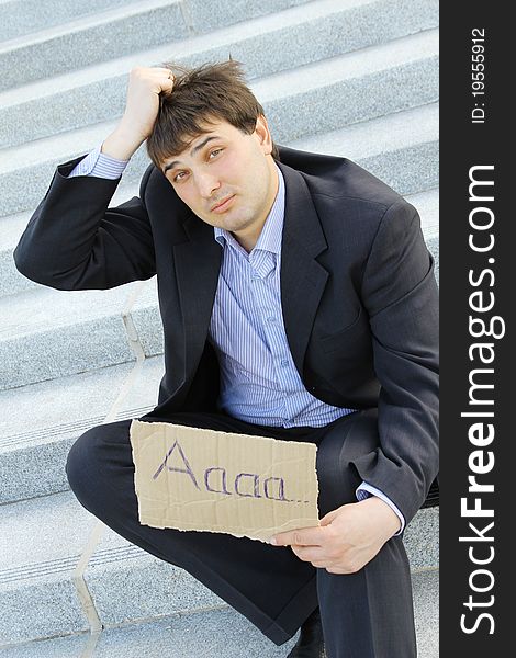 Young businessman sitting on the stairs of the building in hand bumga cardboard with the text aaaa.. with his other hand holding his head. Young businessman sitting on the stairs of the building in hand bumga cardboard with the text aaaa.. with his other hand holding his head