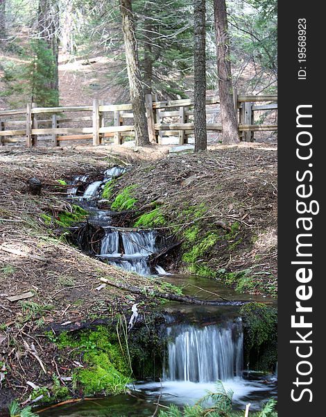 Small stream with waterfalls winding through hills and between trees under wooden fence. Small stream with waterfalls winding through hills and between trees under wooden fence