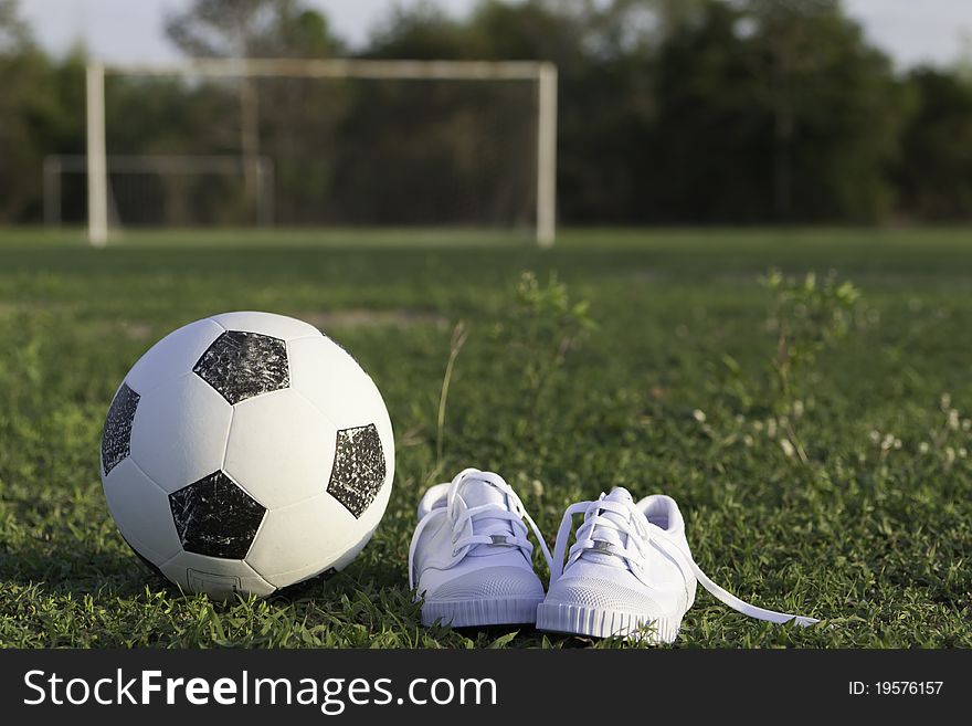 Old ball and white children's gym shoes on a football ground against gate. Old ball and white children's gym shoes on a football ground against gate