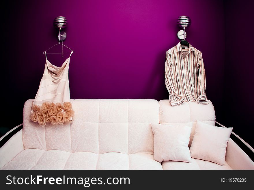 Beautifull white dress of bride and groom's striped shirt are hanging on lamps over a sofa in the beginning of wedding day. Beautifull white dress of bride and groom's striped shirt are hanging on lamps over a sofa in the beginning of wedding day