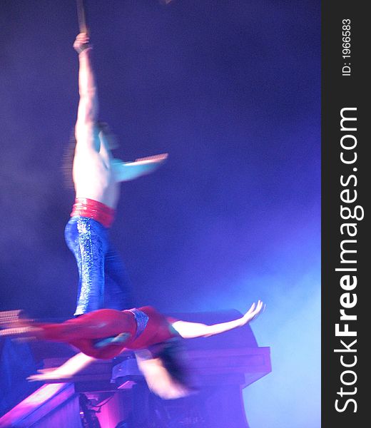 2 aerial acrobats in a spin above the stage, multi-blue/red colors with a misty blue background. Man holding on to rope with one arm as woman is at his feet horizontaly. 2 aerial acrobats in a spin above the stage, multi-blue/red colors with a misty blue background. Man holding on to rope with one arm as woman is at his feet horizontaly.