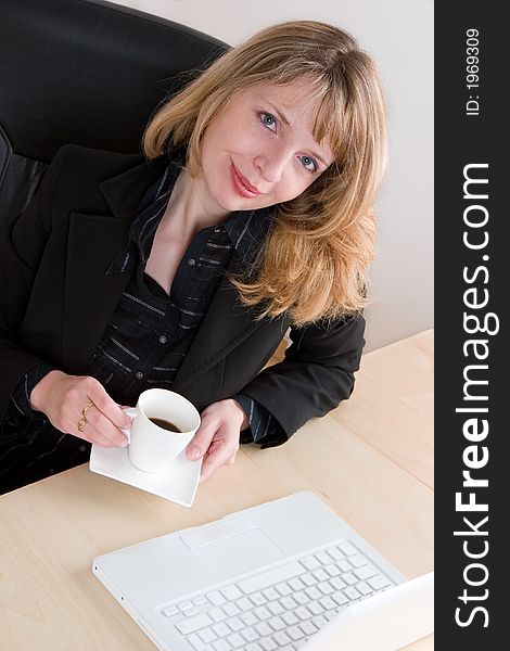 A blond woman in a black suit drinking a cup of coffee at her desk. A blond woman in a black suit drinking a cup of coffee at her desk