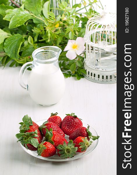 Plate with fresh strawberries and jug of milk on white table with old vintage cage and wild flowers