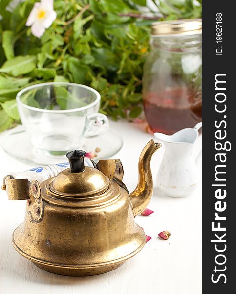 Old golden  teapot on white table with empty transparent teacup and little jug with green leafs as background. Old golden  teapot on white table with empty transparent teacup and little jug with green leafs as background