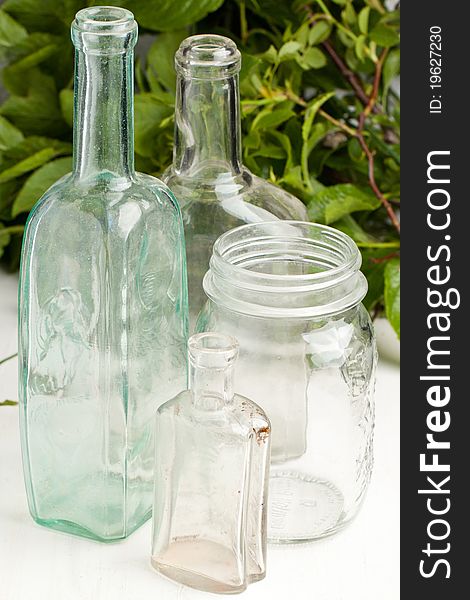 Composition with three vintage bottles and empty pot on white table with green leafs as background