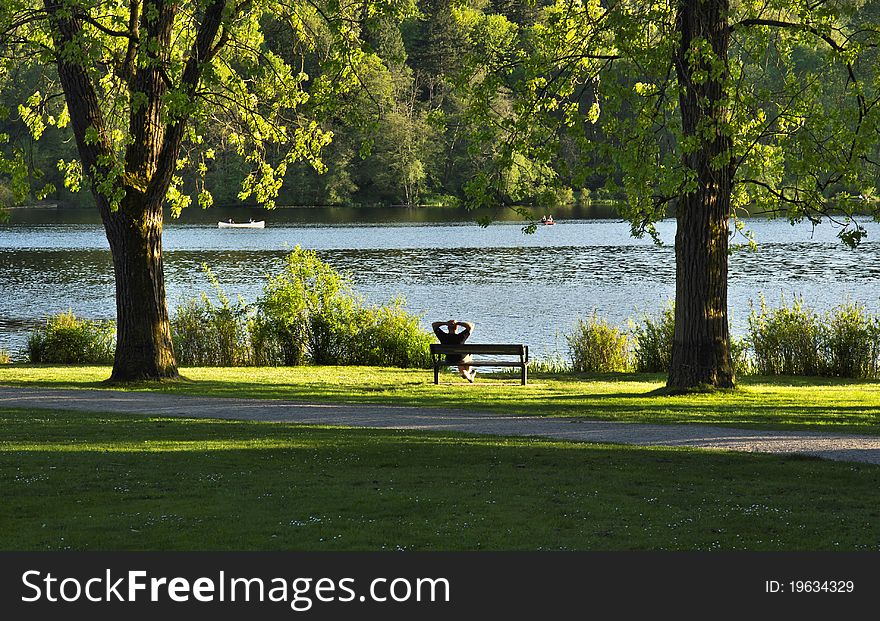 A man relaxes on a park bench as people enjoy a lake. A man relaxes on a park bench as people enjoy a lake