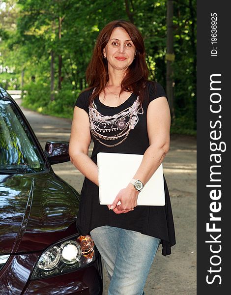 The woman with long hair with the white laptop in hands costs near the car in the street. The woman with long hair with the white laptop in hands costs near the car in the street