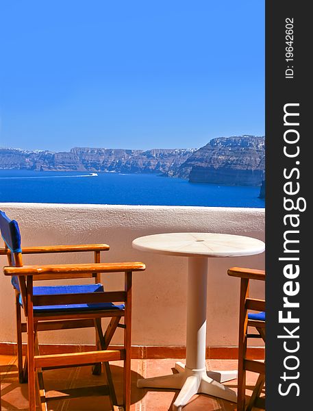 Blue chairs and a small table on beautiful terrace in Santorini with Caldera view and Aegean sea. As always clear blue sky in hot Greece. Great atmosphere for vacation and holidays. Blue chairs and a small table on beautiful terrace in Santorini with Caldera view and Aegean sea. As always clear blue sky in hot Greece. Great atmosphere for vacation and holidays.