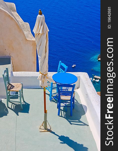 Blue chairs and a small table on beautiful terrace in Santorini with Caldera view and Aegean sea. As always clear blue sky in hot Greece. Great atmosphere for vacation and holidays. Blue chairs and a small table on beautiful terrace in Santorini with Caldera view and Aegean sea. As always clear blue sky in hot Greece. Great atmosphere for vacation and holidays.