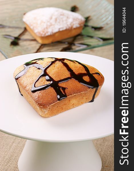 A small heart-shaped cake with chocolate topping served on a cake stand. A small cake with icing sugar served on a glass plate out of focus in the background. A small heart-shaped cake with chocolate topping served on a cake stand. A small cake with icing sugar served on a glass plate out of focus in the background