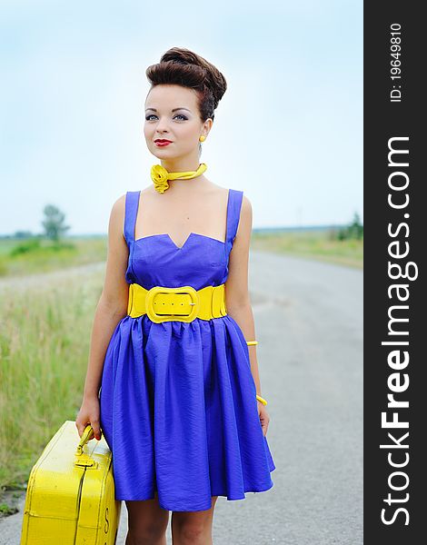 Beautiful smiling girl in a blue dress with a yellow suitcase on the road