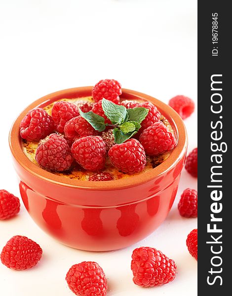 French creme brulee dessert with raspberries and mint covered with caramelized sugar in red terracotta ramekin on white background