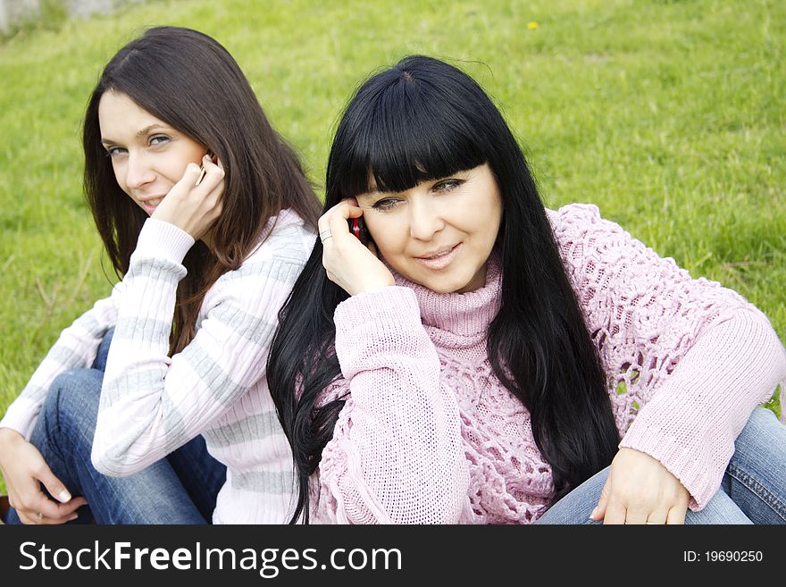Attractive mother and teenage daughter talk on the phone outdoors in a park sitting back to back. In the foreground a mother. Attractive mother and teenage daughter talk on the phone outdoors in a park sitting back to back. In the foreground a mother