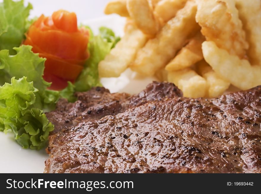 A horizontal close-up image of a portion of steak served on a white ceramic plate with french fries, lettuce and a tomato. A horizontal close-up image of a portion of steak served on a white ceramic plate with french fries, lettuce and a tomato