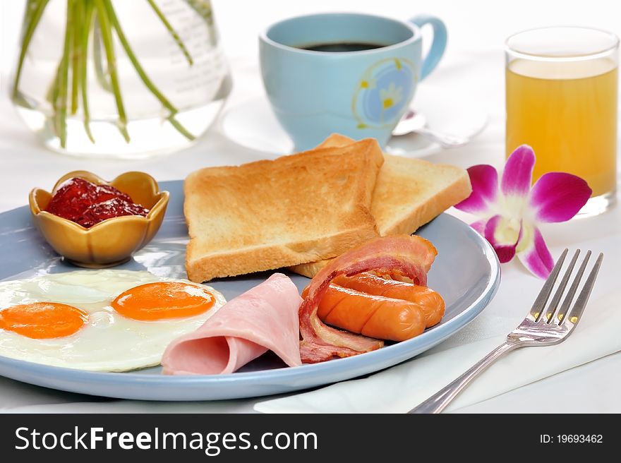 Eggs, bacon and sausages served with toast with strawberry jam served on a blue ceramic plate accompanied by coffee, pineapple juice and an orchid. Eggs, bacon and sausages served with toast with strawberry jam served on a blue ceramic plate accompanied by coffee, pineapple juice and an orchid