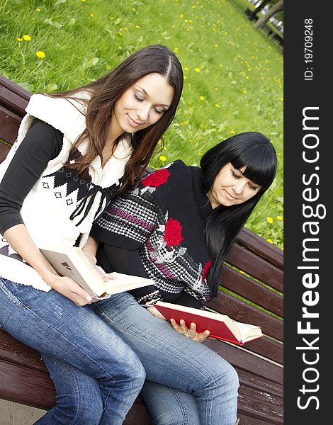 Attractive Mom and daughter, a teenager in a park reading a book and make merry while sitting on a wooden bench. Attractive Mom and daughter, a teenager in a park reading a book and make merry while sitting on a wooden bench