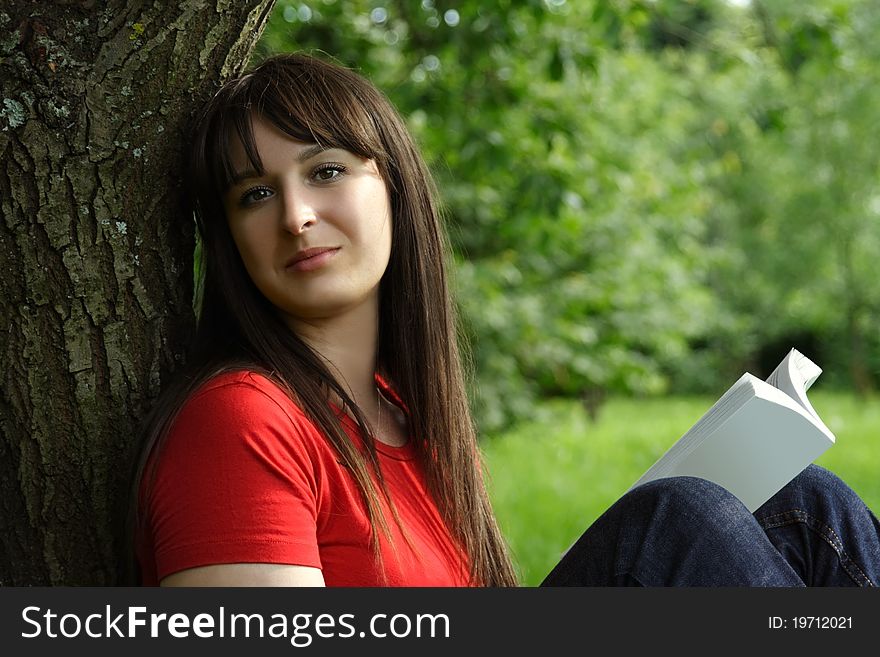 Young brunette girl in red shirt siting near tree and reading book, outdoor. Young brunette girl in red shirt siting near tree and reading book, outdoor