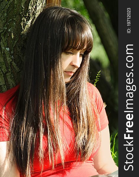 Young brunette girl in red shirt siting near tree