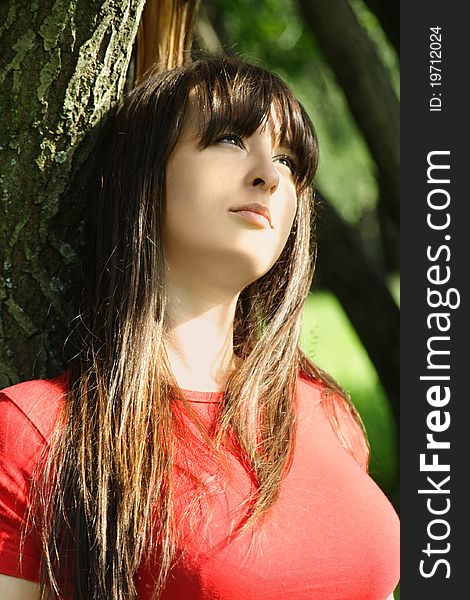 Young brunette girl in red shirt sitting near tree and looking at side. Young brunette girl in red shirt sitting near tree and looking at side