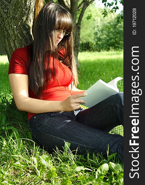 Young brunette girl in glasses and red shirt sitting near tree and reading a book. Young brunette girl in glasses and red shirt sitting near tree and reading a book