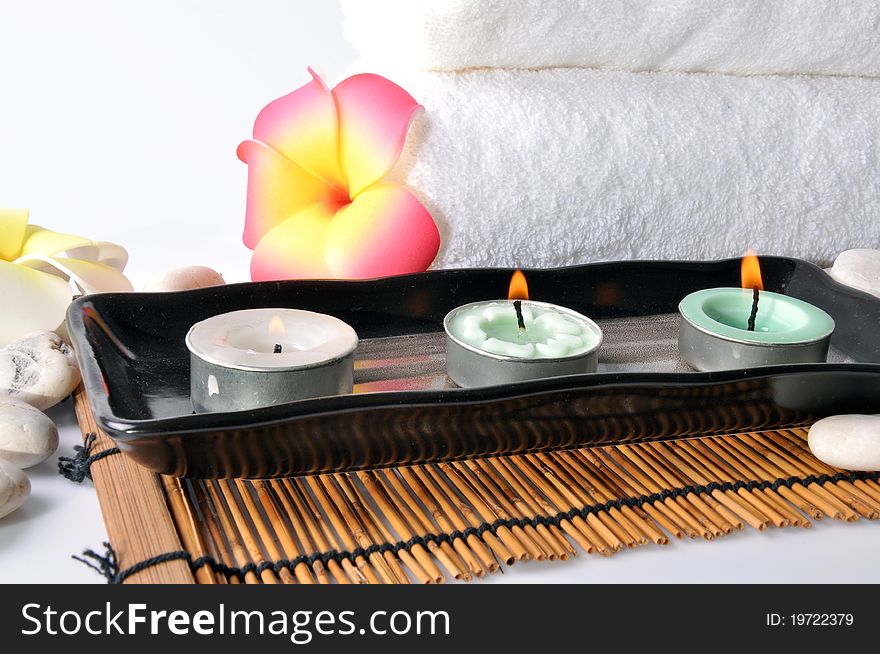 Three candles in plate with white towel and plumeria on bamboo mat.