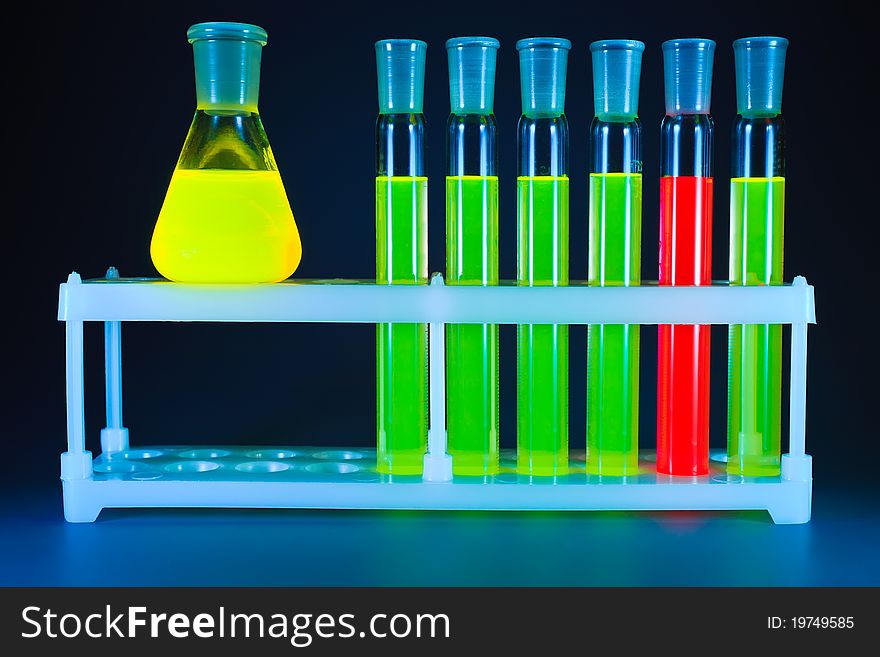 Five test tubes with a green fluorescing liquid and one with a red liquid in a support and yellow flask on a dark blue background. Five test tubes with a green fluorescing liquid and one with a red liquid in a support and yellow flask on a dark blue background.