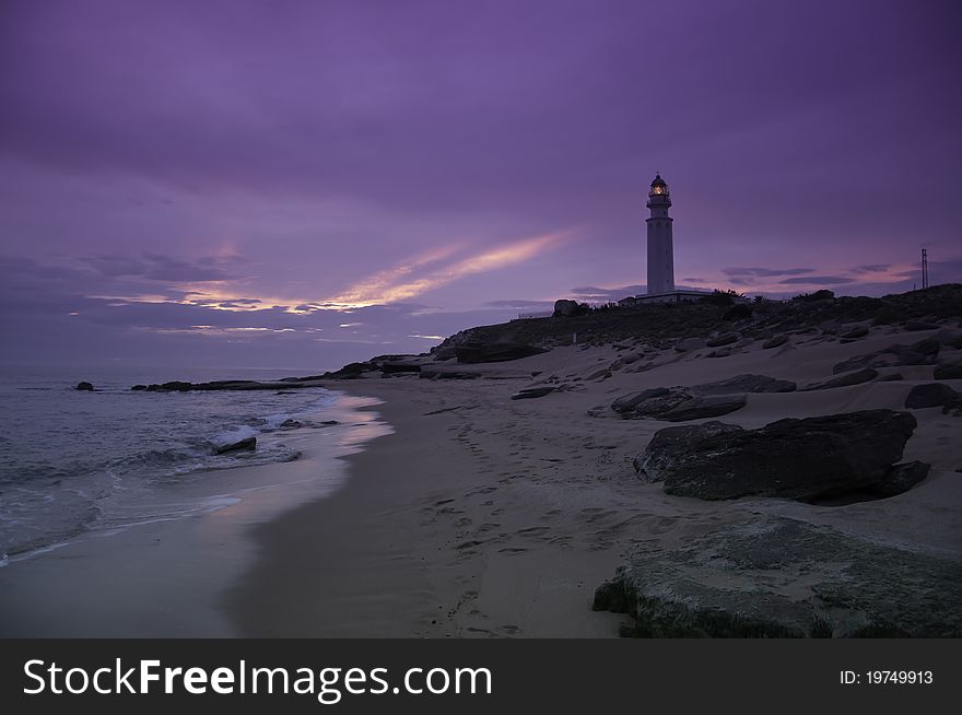 A tranquil beach with a purple sunset with a lighthouse in the background