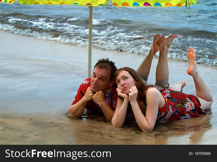 A couple vacationing in Hawaii seem to be bored with paradise as they lie under an umbrella at the shore with the ocean waves at their feet. A couple vacationing in Hawaii seem to be bored with paradise as they lie under an umbrella at the shore with the ocean waves at their feet.