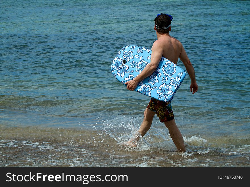 Man walks along the shore of a beach holding a bodyboard, looking for waves. He splashes water as he walks. Man walks along the shore of a beach holding a bodyboard, looking for waves. He splashes water as he walks.