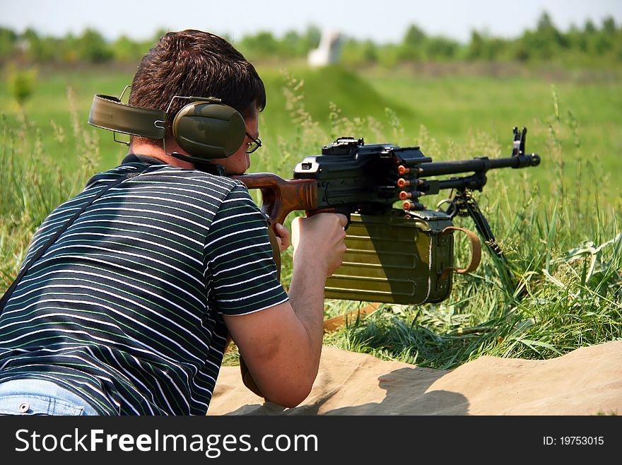 A man in a dark shirt and headphones lies with a machine gun and shoots. A man in a dark shirt and headphones lies with a machine gun and shoots