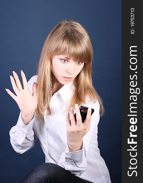 The young beautiful girl-blonde looks at phone with surprise on a dark blue background. The young beautiful girl-blonde looks at phone with surprise on a dark blue background