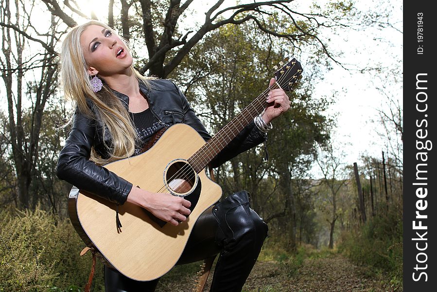 A beautiful blonde young lady is playing a guitar and singing in the forest, she could also be worshipping. A beautiful blonde young lady is playing a guitar and singing in the forest, she could also be worshipping