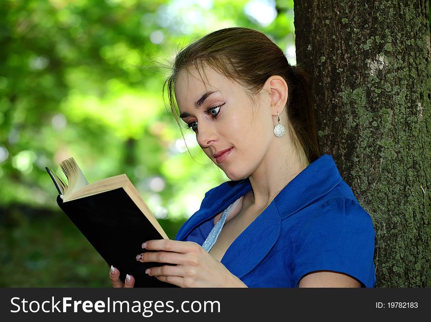 The beautiful young girl the teenager reads the book in park, having leaned against a tree. The beautiful young girl the teenager reads the book in park, having leaned against a tree