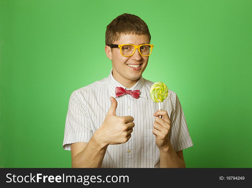 Closeup of an attractive young man in a shirt, bow tie and yellow sunglasses with a big lollipop. Thumbs up. Closeup of an attractive young man in a shirt, bow tie and yellow sunglasses with a big lollipop. Thumbs up