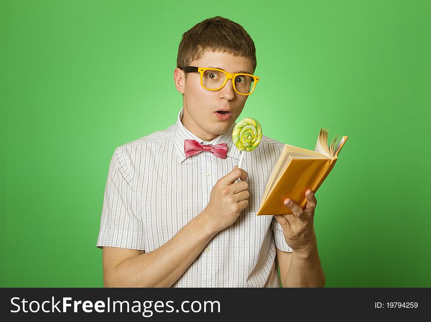 Young attractive man in a shirt and yellow glasses, bow tie on a green background reads the yellow book and a round lollipop. Bookworm. Young attractive man in a shirt and yellow glasses, bow tie on a green background reads the yellow book and a round lollipop. Bookworm