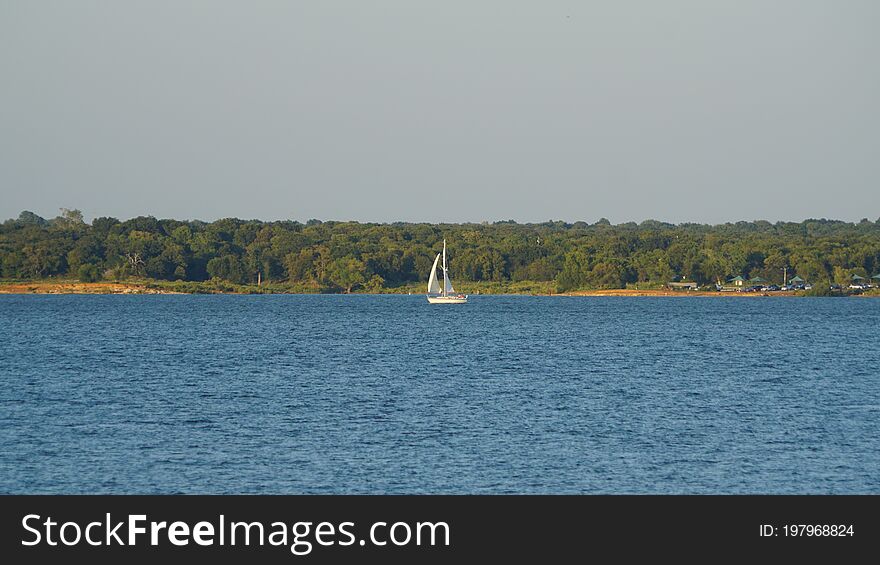 A photograph of one lone white colored sailboat sailing on the Grapevine Lake in Texas. A photograph of one lone white colored sailboat sailing on the Grapevine Lake in Texas.