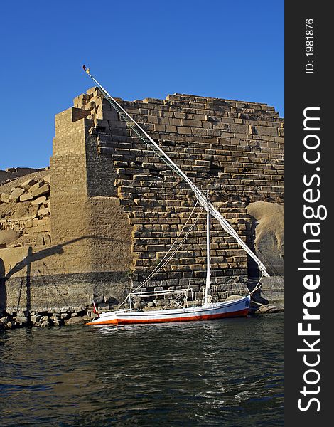 Anchored Egyptian traditional sailing boat know as Felucca shot against a beautiful blue sky on a sunny winter day in Aswan. The background is an ancient building with some phraonic scripts on some of its stones. Anchored Egyptian traditional sailing boat know as Felucca shot against a beautiful blue sky on a sunny winter day in Aswan. The background is an ancient building with some phraonic scripts on some of its stones.