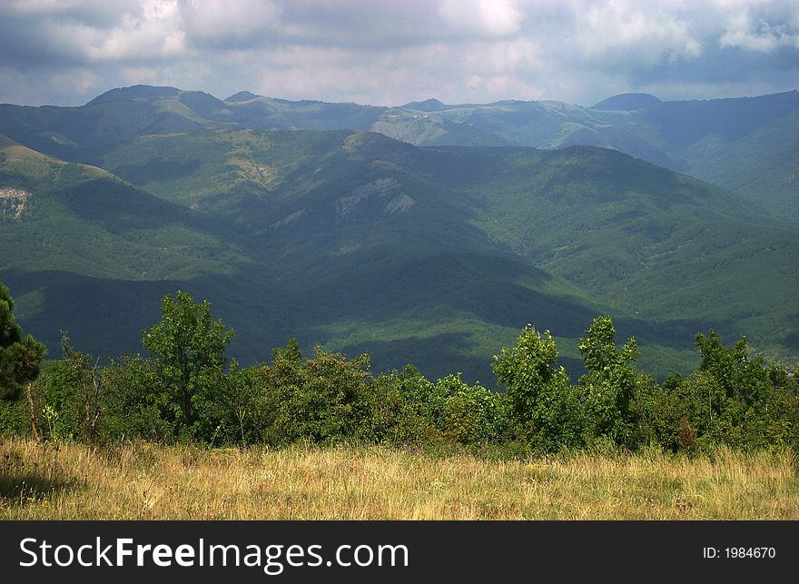 The image of mountains with shadows from clouds and a glade with a dry grass and green bushes. The image of mountains with shadows from clouds and a glade with a dry grass and green bushes