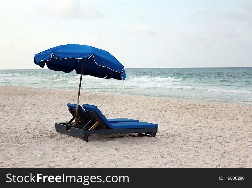 A lone pair of blue beach lounge chairs shaded by a matching umbrella with a view of the waves breaking on the shore. Horizontal shot. A lone pair of blue beach lounge chairs shaded by a matching umbrella with a view of the waves breaking on the shore. Horizontal shot.