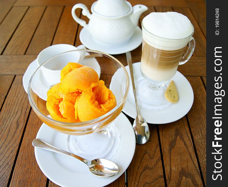 Orange ice cream in a glass bowl and glass of coffee latte  on the background of the wooden table surface. Orange ice cream in a glass bowl and glass of coffee latte  on the background of the wooden table surface