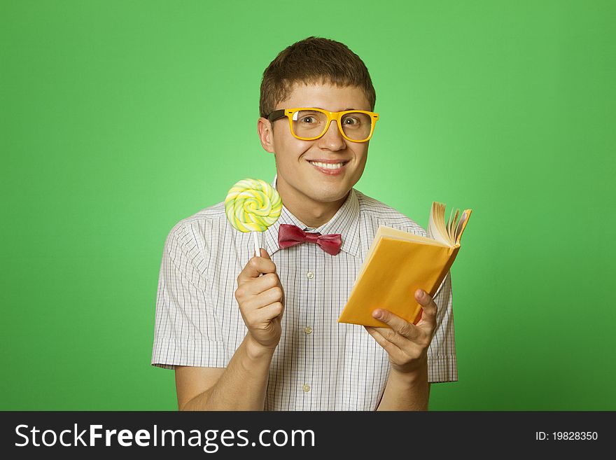 Young attractive man in a shirt and yellow glasses, bow tie on a green background reads the yellow book and a round lollipop. Bookworm. Young attractive man in a shirt and yellow glasses, bow tie on a green background reads the yellow book and a round lollipop. Bookworm