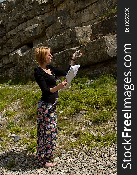 A young woman in floral pants and black top holding a clipboard inspects a rock specimen infront of a limestone cliff. A young woman in floral pants and black top holding a clipboard inspects a rock specimen infront of a limestone cliff.