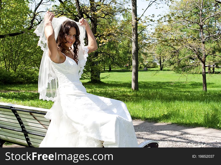 Beautiful young bride sitting on a park bench, smiling and holding veil