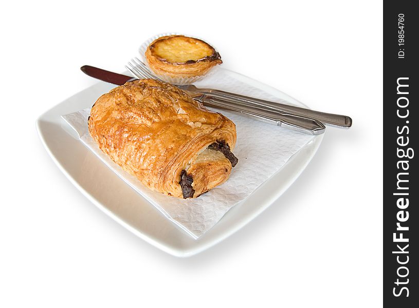 A croissant filled with chocolate and a Pastel de Nata on a plate with knife and fork. Isolated with copy space. A croissant filled with chocolate and a Pastel de Nata on a plate with knife and fork. Isolated with copy space.