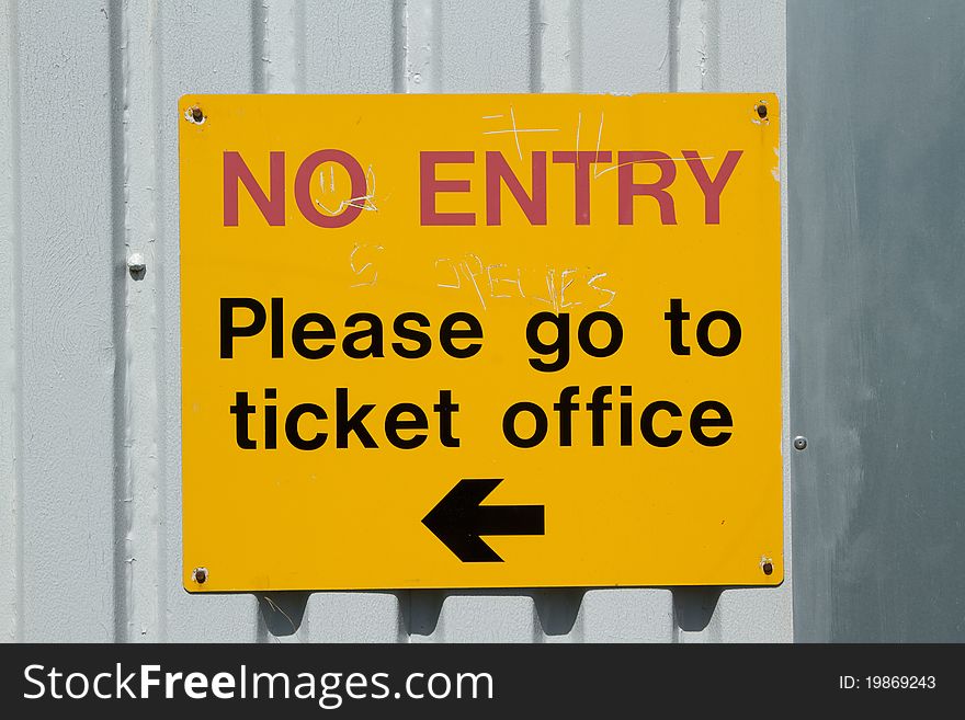 A yellow sign against a grey metal background with the words 'no entry please go to ticket office' with a directional arrow.