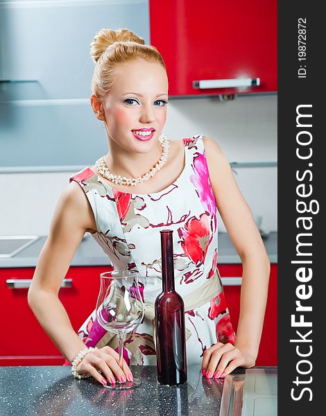 Young beautiful blonde girl wearing white and pink dress holding glass and bottle with wine in interior of modern red kitchen in expectation of party. Young beautiful blonde girl wearing white and pink dress holding glass and bottle with wine in interior of modern red kitchen in expectation of party