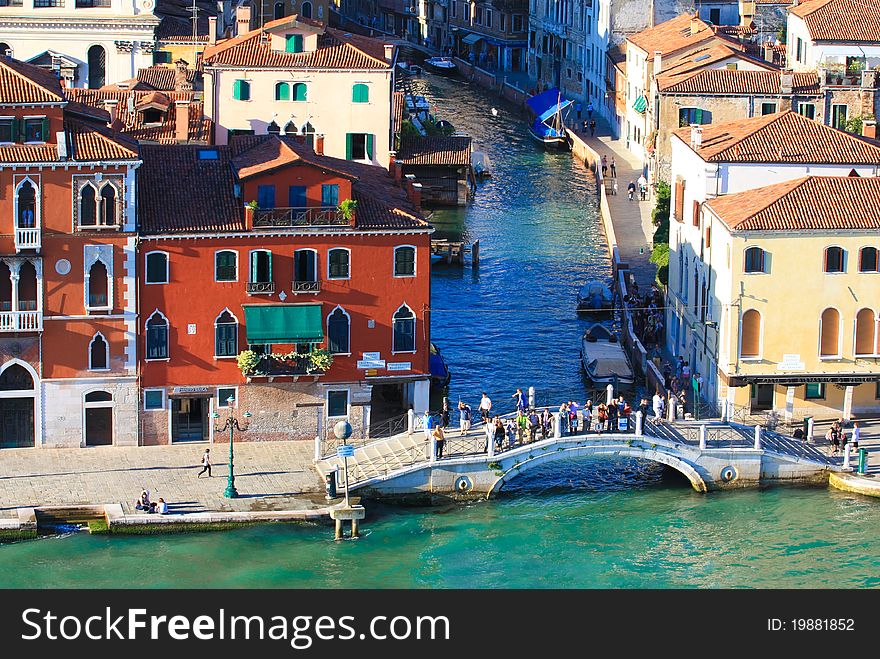 The view from atop of the straight canal of Venice with houses and boats lined up along and people on the bridge. The view from atop of the straight canal of Venice with houses and boats lined up along and people on the bridge